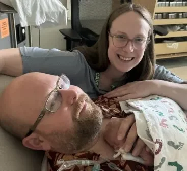 A smiling woman with glasses leans over a man with a beard and glasses lying down. A tiny infant rests on his chest under a footprint-patterned blanket, connected to medical tubing in a clinical hospital setting.