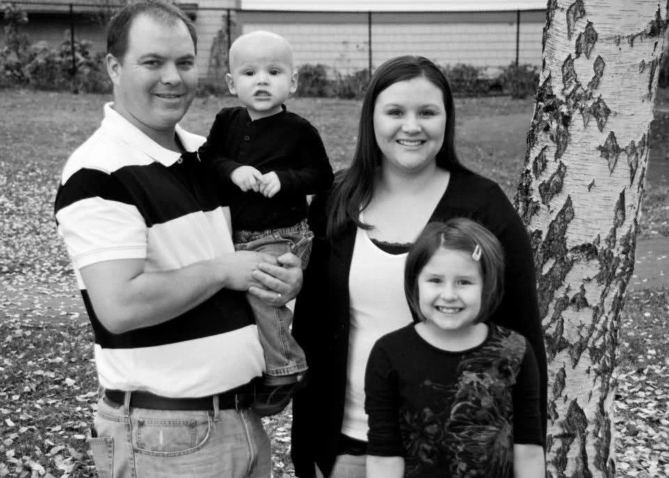 Black and white photo of Kyler as a baby held by dad with mom and older sister outside next to a tree