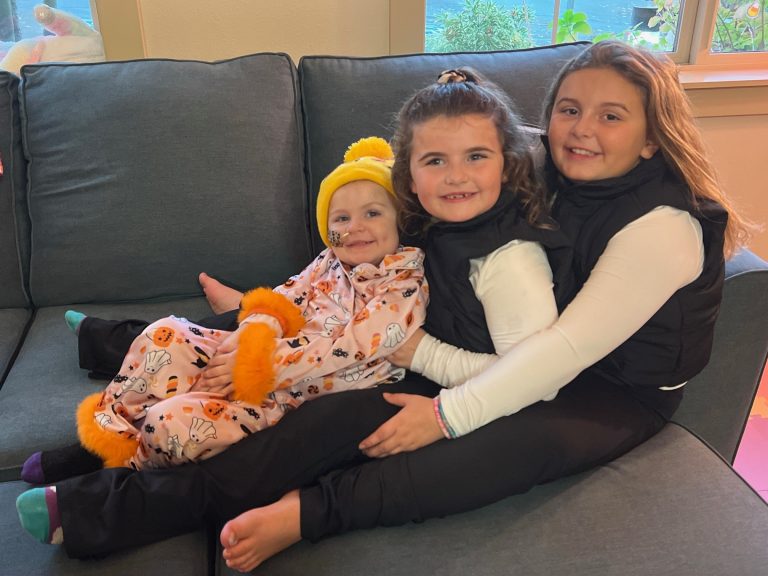 Three sisters ages 3, 6, and 8 sitting in each other's laps, on a gray couch, smiling.