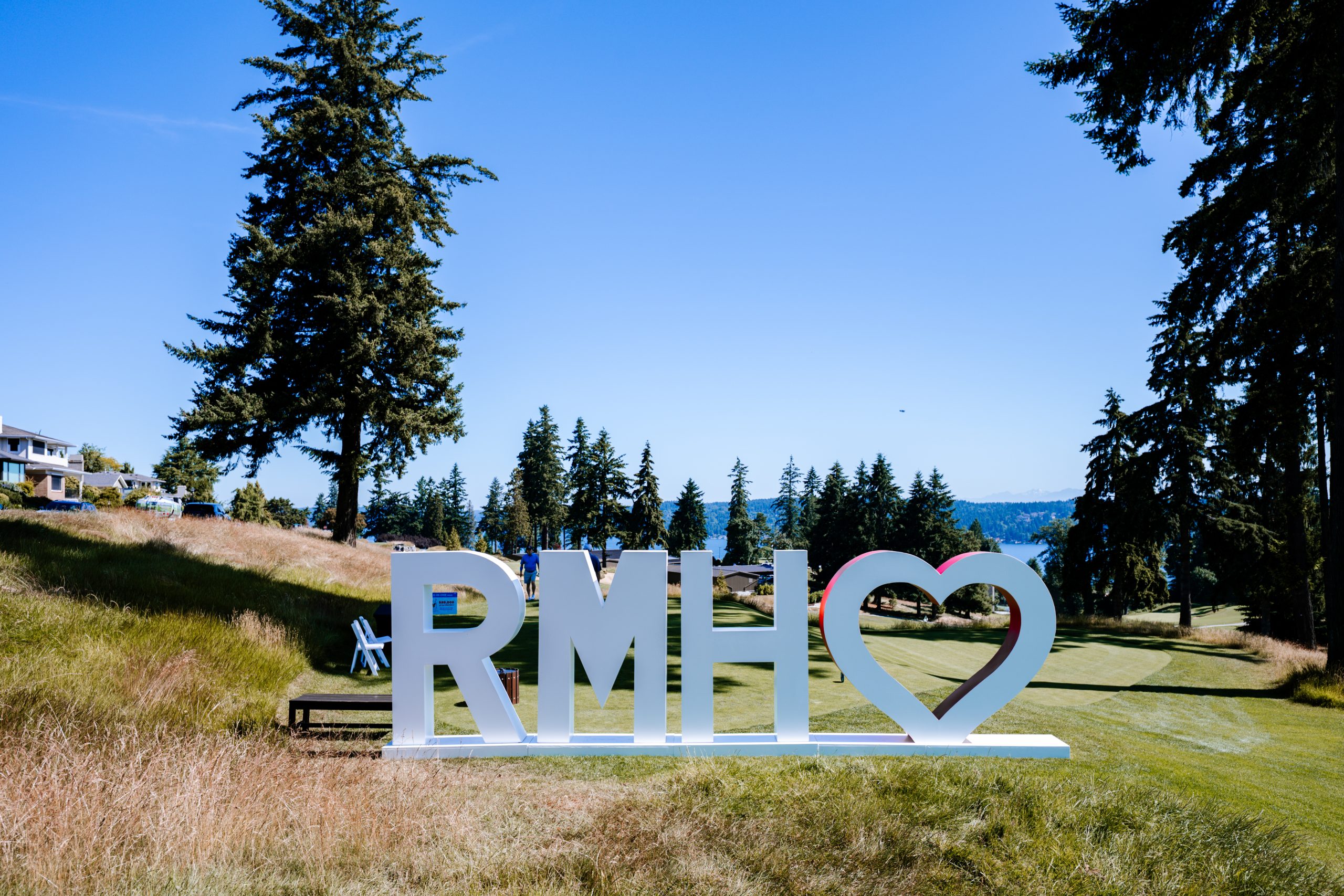 Large white "RMH" letters with a heart symbol stand on a golf course tee box during a fundraising tournament. Towering evergreen trees frame the scene, with rolling fairways, a distant lake, and blue skies.