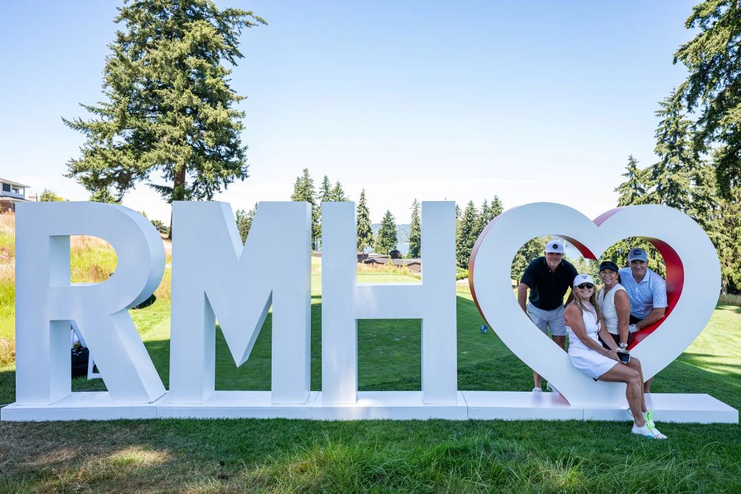 Four participants in golf attire smile and pose inside a large, white RMH & heart installation on a sunny golf course. The massive white letters spell out "RMH" followed by the heart symbol against a backdrop of green grass and trees.