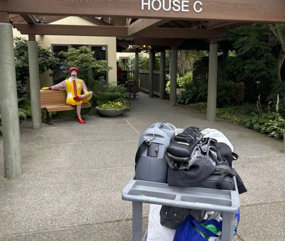 A grey utility cart loaded with bags stands on a concrete pathway. In the background, a wooden structure is labeled "HOUSE C" across the beam. To the left, a Ronald McDonald statue is seated on a wooden bench surrounded by green plants.