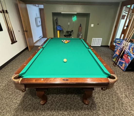 A large wooden pool table with green felt sits in the center of a carpeted room. A single yellow ball rests near the center of the table, while a racked triangle of billiard balls sits at the far end under a small green light.