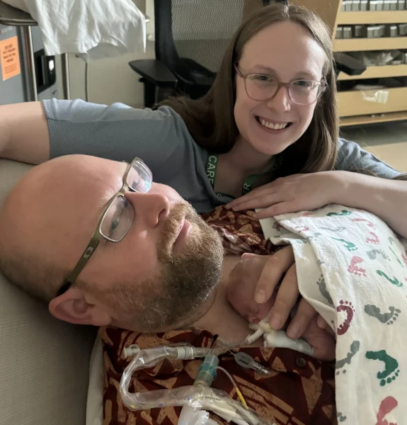 A smiling woman with glasses leans over a man with a beard and glasses lying down. A tiny infant rests on his chest under a footprint-patterned blanket, connected to medical tubing in a clinical hospital setting.